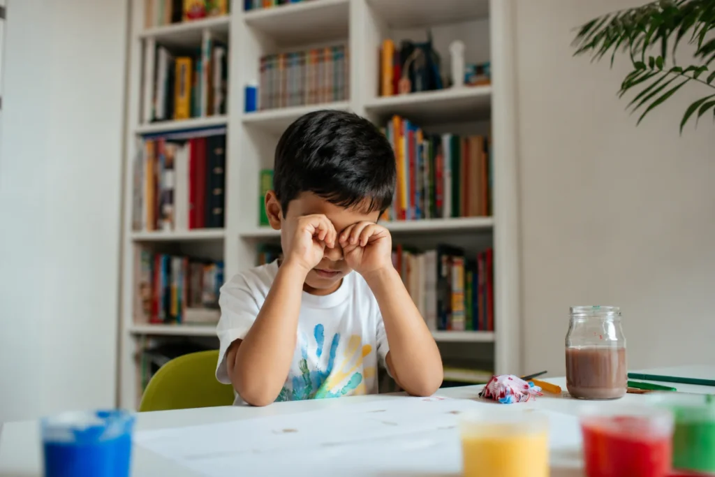 Tired child rubbing eyes while sitting at a table, illustrating how poor sleep, lack of movement, and physical fatigue can affect focus and concentration in children.